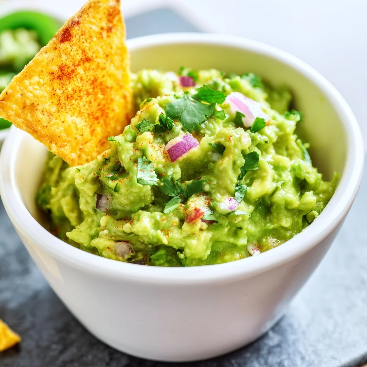 A generous scoop of chunky Big Game Guacamole rests on a crispy homemade tortilla chip, showcasing ripe avocado, red onion, and jalapeño against a dark, textured background.