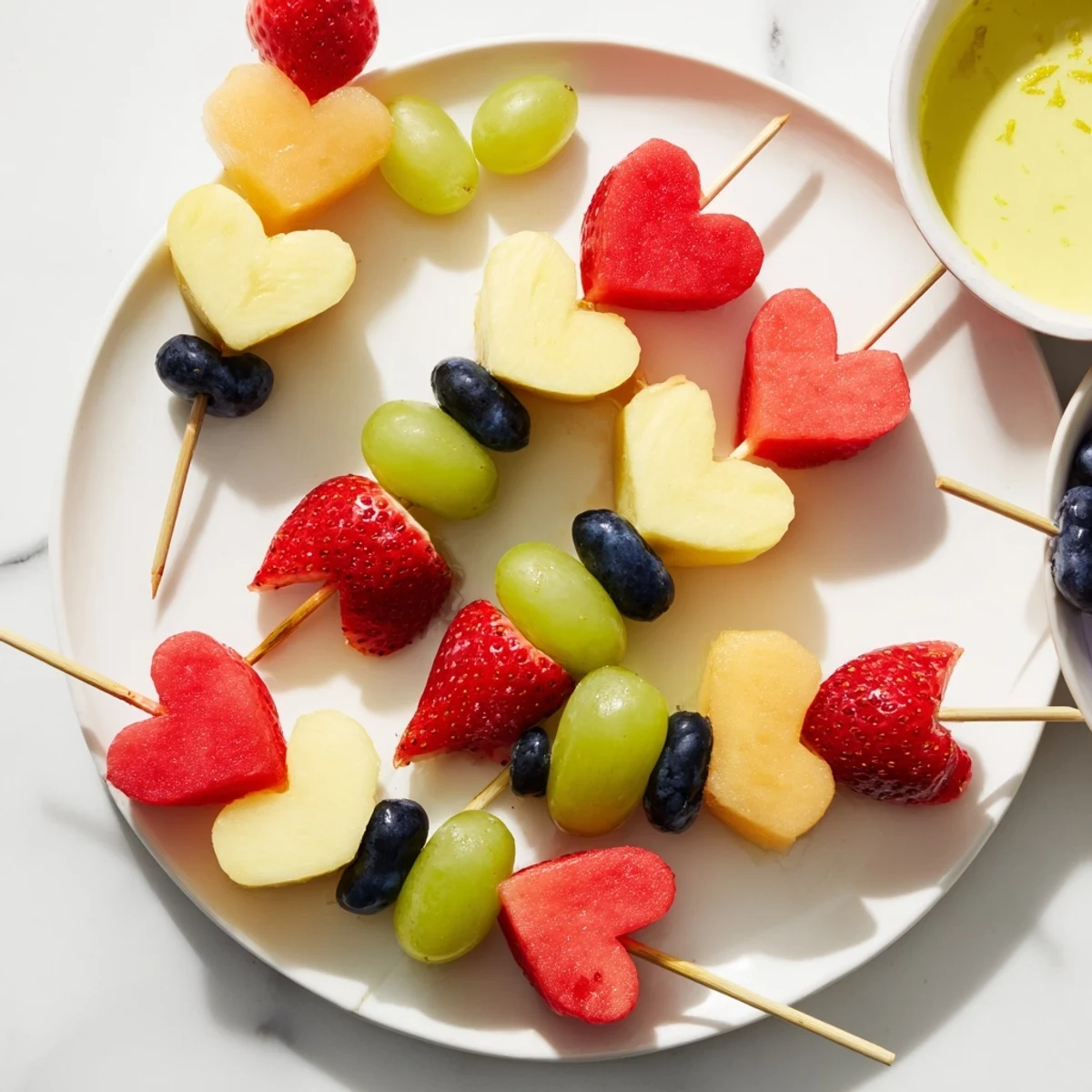 Colorful heart-shaped fruit skewers with strawberries, watermelon, and grapes on wooden sticks, served with creamy honey-yogurt dip for a healthy snack.