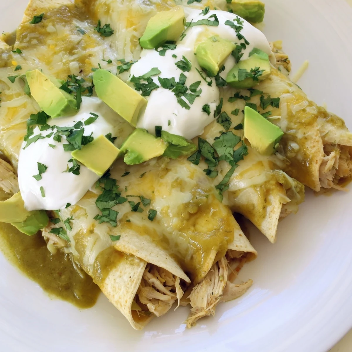 A close-up shot shows freshly baked Green Enchiladas With Chicken served on a plate, topped with creamy sour cream, diced avocado, and cilantro.