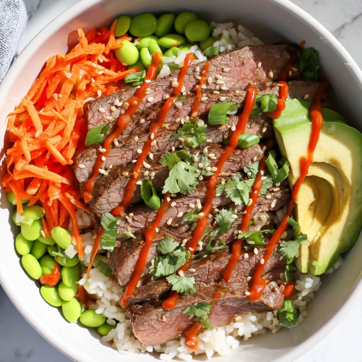 Protein-packed flank steak bowl featuring fresh avocado, edamame, carrots, and zesty sriracha sauce