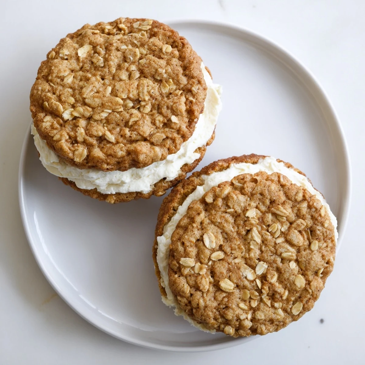 Golden brown oatmeal cookies sandwiching sweet white cream filling on a white serving plate