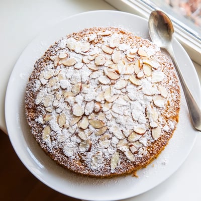 A slice of moist Almond Cake on a white plate, showing tender crumb, topped with berries and a dollop of whipped cream.